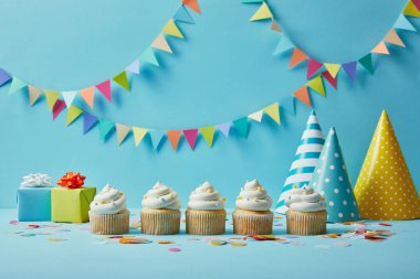 Şeker sprinkles, parti şapkaları ve renkli kiraz kuşu ile mavi arka plan üzerinde hediyeler ile lezzetli cupcakes