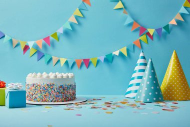 Tasty cake with sugar sprinkles,party hats and gifts on blue background with colorful bunting clipart