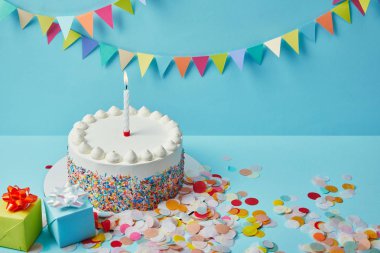 Delicious cake with sugar sprinkles, gifts and confetti on blue background with colorful bunting clipart