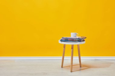 Little wooden table with pile of magazines and white cup of coffee near yellow wall clipart