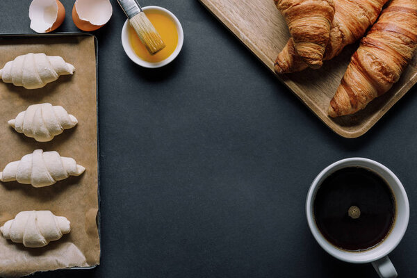 top view of dough for croissants on tray, egg yolk with brush and coffee cup on black surface