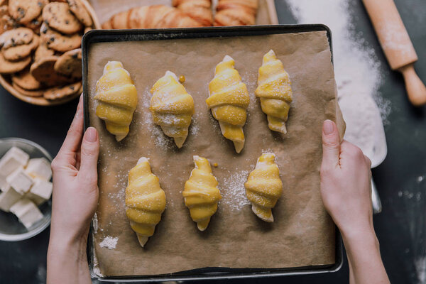 cropped image of woman holding tray with dough for croissants over table ingredients 