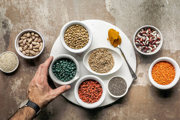 cropped view of man holding spirulina with superfoods and legumes on textured rustic background