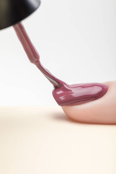 purple nail polish on fingernail of woman with wet brush on grey background