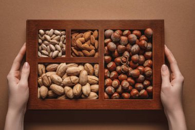 partial view of woman holding box with assortment of different nuts on brown background clipart