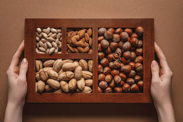 partial view of woman holding box with assortment of different nuts on brown background