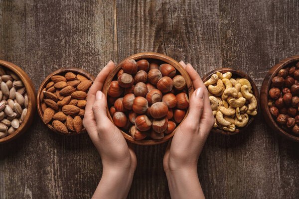 partial view of woman holding bowl with hazelnuts on wooden tabletop with different nuts around