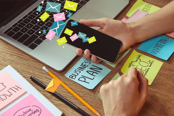 cropped image of woman holding smartphone with email icons at table with business plan sticker