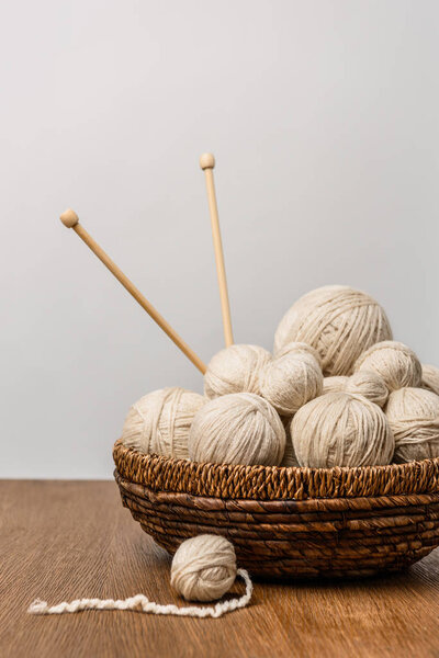 close up view of knitting balls with knitting needles in wicker basket on wooden surface on grey backdrop