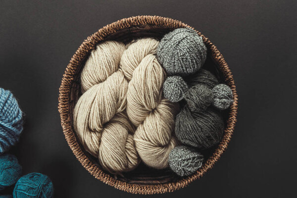 top view of beige and grey knitting clews in wicker basket on grey backdrop with blue yarn