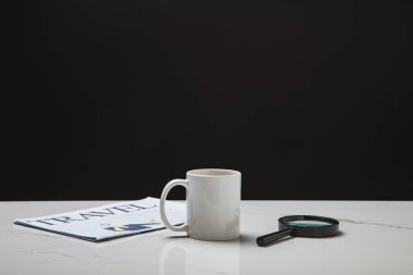 close-up view of cup, magnifying glass and travel newspaper on white surface on black clipart