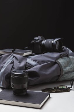 backpack, photo camera with lens, notebooks with pen and compass on black   clipart