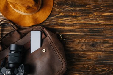 ahşap masa üzerinde kahverengi deri çanta, şapka, fotoğraf makinesi ve smartphone üstten görünüm