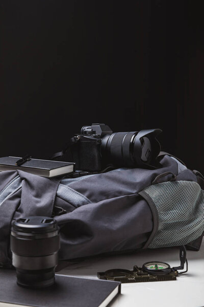 backpack, photo camera with lens, notebooks with pen and compass on black, travel concept
