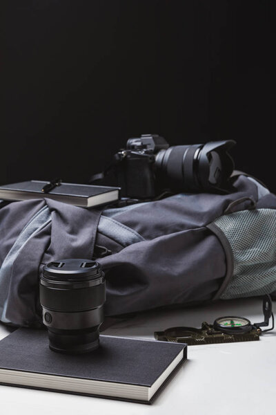 backpack, photo camera with lens, notebooks with pen and compass on black  