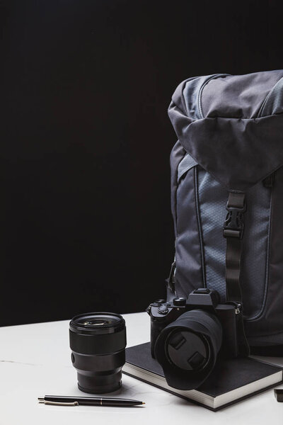 backpack, photo camera with lens and notebook with pen on black 