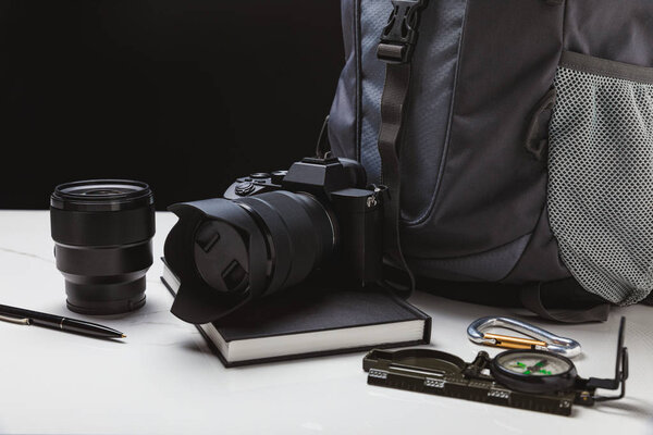 close up view of photo camera with lens, pen, notebook, compass and backpack on black