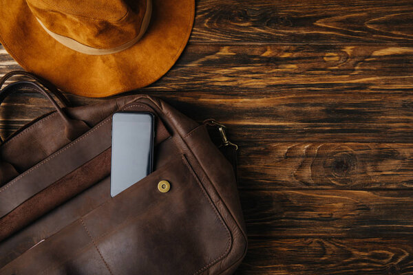 top view of brown leather bag with smartphone and hat on wooden table, travel concept