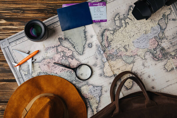 top view of brown leather bag, hat, magnifying glass, small model plane, photo camera, lens and passport with boarding pass on map 