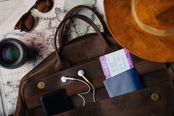 вид сверху кожаная сумка с паспортом, билет, смартфон с наушниками, солнцезащитные очки, объектив и шляпа на карте
