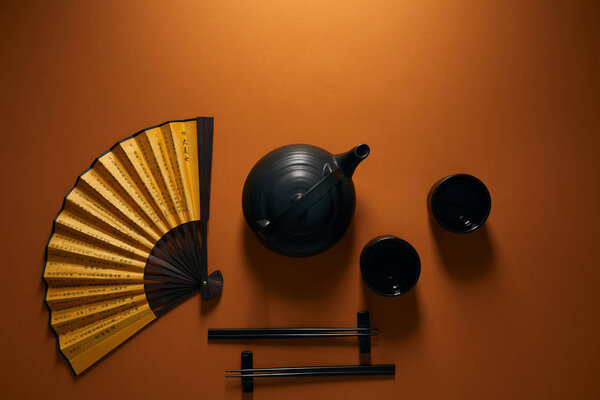 top view of black tea set, chopsticks and golden fan with hieroglyphs on brown