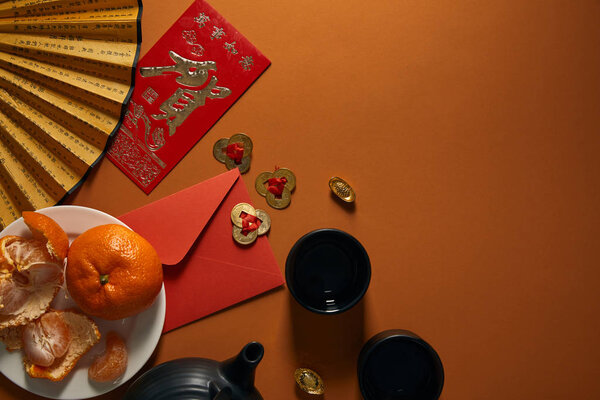 top view of tangerines on plate, black tea set, fan with hieroglyphs, golden decorations and red envelope on brown background