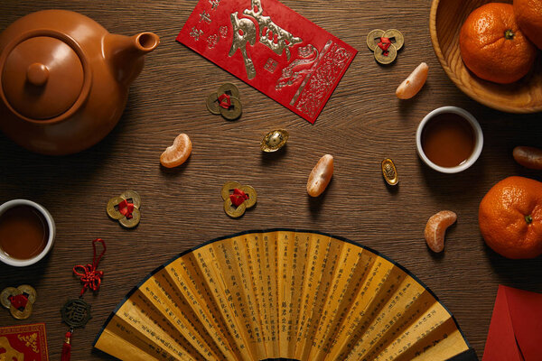 top view of fresh ripe tangerines, tea set, fan with hieroglyphs and golden coins on wooden table 
