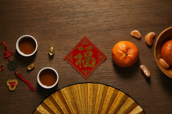 fan, fresh ripe tangerines, golden hieroglyph, cups of tea and coins on wooden surface 