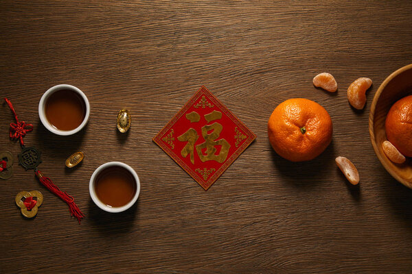top view of fresh ripe tangerines, golden hieroglyph, cups of tea and coins on wooden surface 