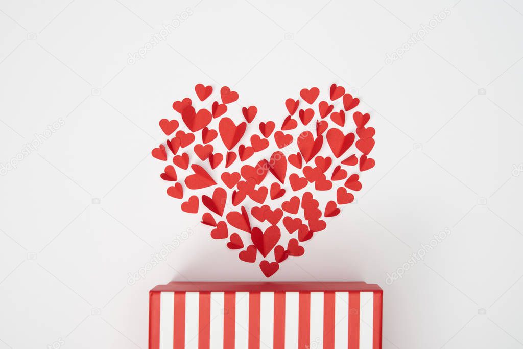 top view of heart shaped arrangement of small red paper cut hearts and striped gift box on white background