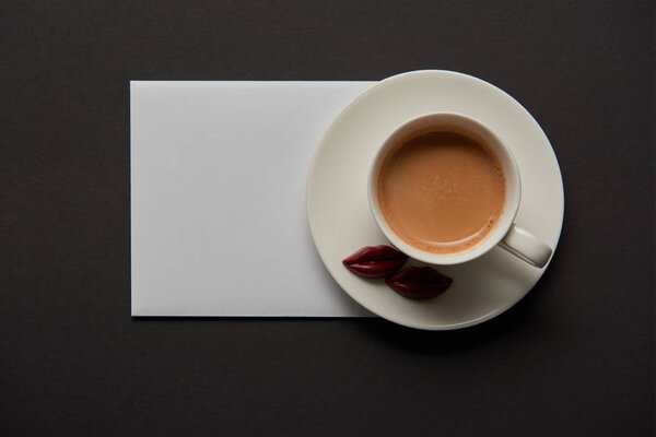 top view of cup with coffee, chocolate lips on saucer and empty white card on black background