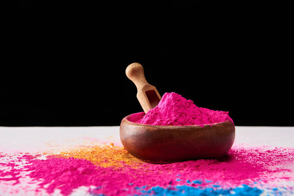 wooden spatula and bowl with pink holi powder on white table isolated on black