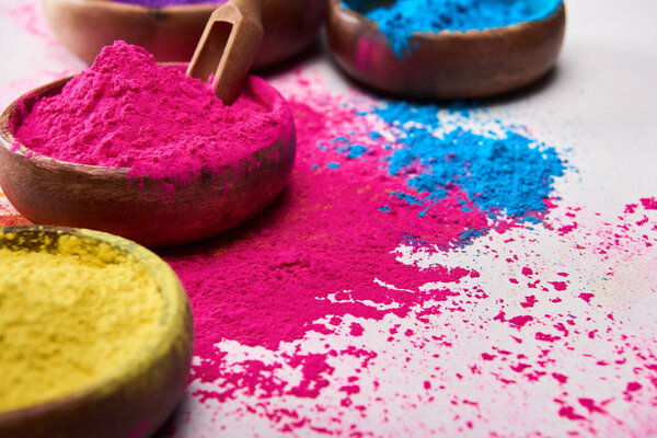 wooden spatula and bowls with pink, blue and yellow holi powder on white background