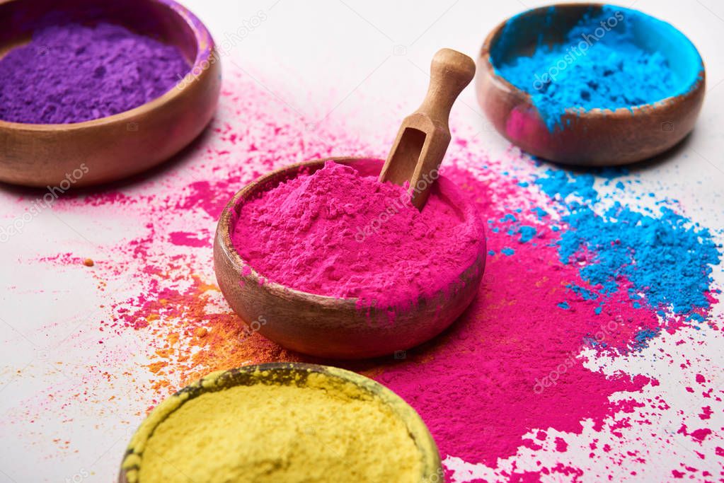 wooden spatula and bowls with yellow, pink, blue and purple holi powder on white background