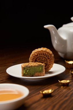 selective focus of traditional chinese mooncakes, tea pot and gold ingots on wooden table isolated on black clipart