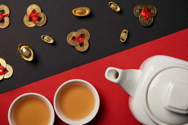 top view of tea pot, cups, gold ingots and feng shui coins on red and black background 
