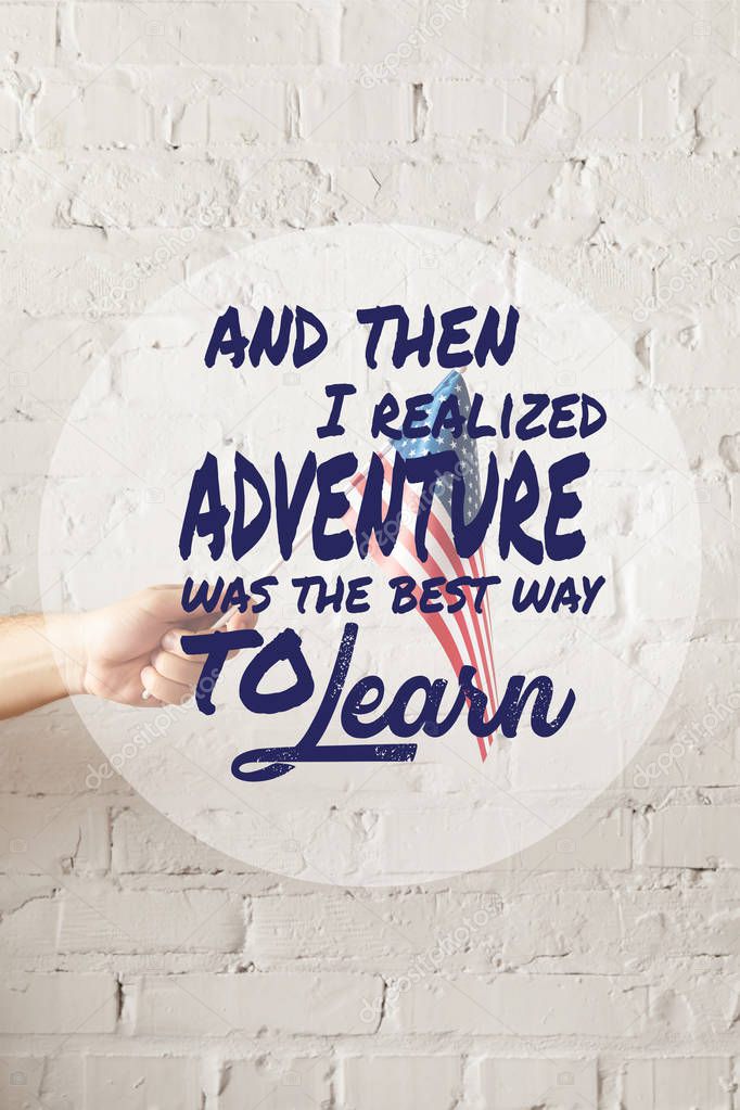 cropped image of man holding american flagpole against white brick wall with and then i realized adventure was the best way to learn quote 