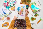 pohled shora ženy držící kreslení na kolena a malování akvarelů, v ní maluje obklopeni barevnými obrázky