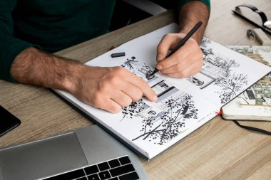 selective focus of mans hands drawing in album on wooden table next to laptop clipart