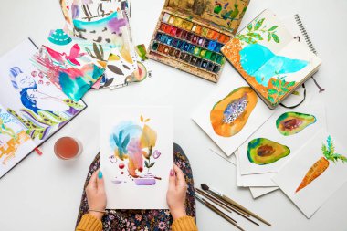 top view of woman holding watercolor drawing while surrounded by color pictures clipart