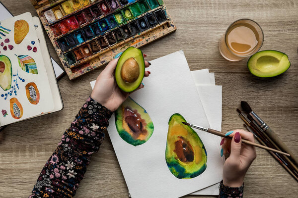 вид женских рук, рисующих авокадо с акварельными красками и кистью на деревянном столе
