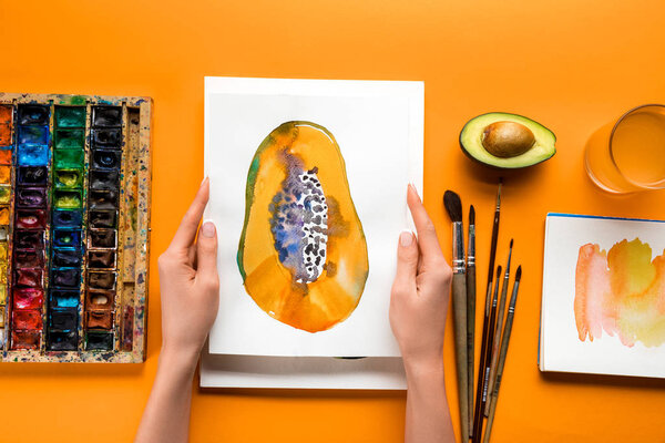 top view of female hands holding papaya picture over yellow table with drawing utensils