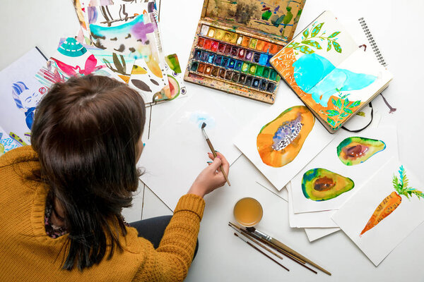 top view of woman painting with watercolors paints while surrounded by color drawings and drawing utensils