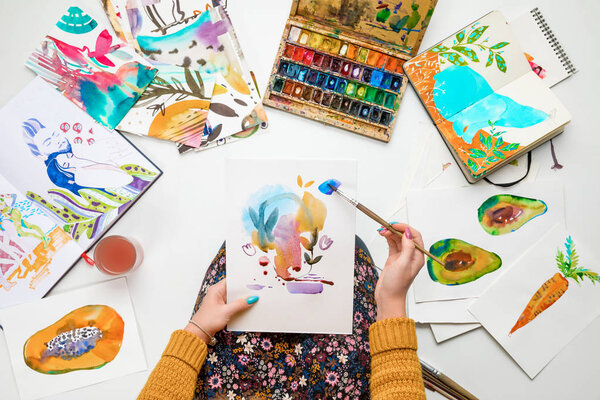 top view of woman holding drawing on knees and painting in it with watercolors paints while surrounded by colored pictures