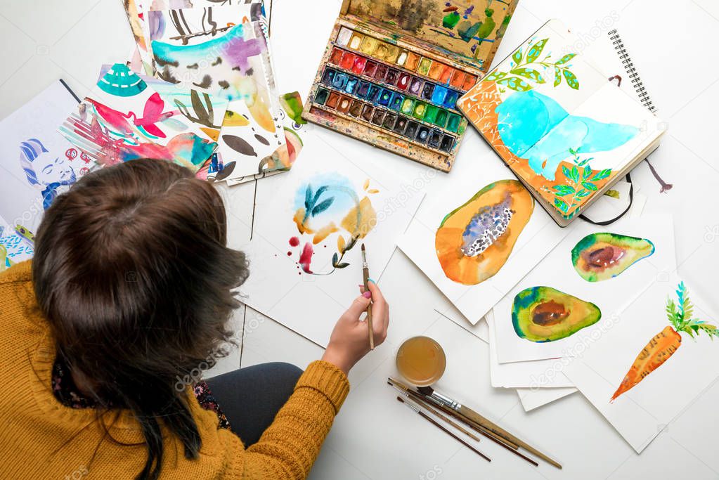 top view of woman painting with watercolors paints while surrounded by color drawings and drawing utensils