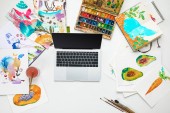 top view of laptop surrounded by watercolors paints drawings