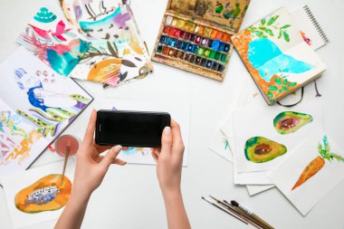 selective focus of female hands making photo of watercolor paintings and drawing utensils using smartphone clipart
