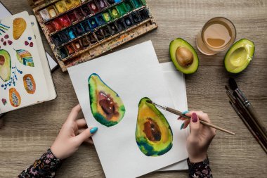 top view of female hands drawing avocados with watercolor paints and paintbrush on wooden table clipart