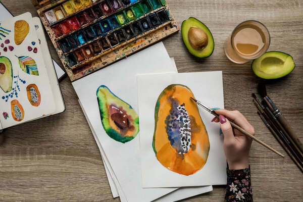 top view of female hands drawing avocado and papaya with watercolor paints, paintbrush on wooden table