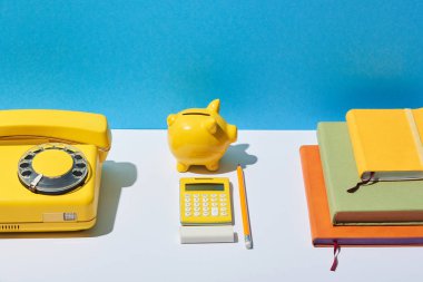 multicolored notebooks, calculator, telephone and piggy bank on white desk and blue background clipart